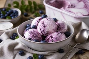 Cremiges Heidelbeer-Joghurt-Eis in einer hellgrauen Schüssel, garniert mit frischen Heidelbeeren und einem Zweig Minze, auf einer Holzunterlage