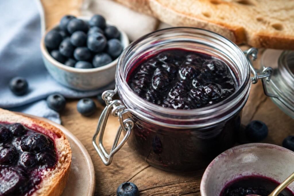 Selbstgemachte Heidelbeermarmelade ohne Gelierzucker in einem Einmachglas, daneben frische Heidelbeeren und ein Holzlöffel auf einem Holzbrett