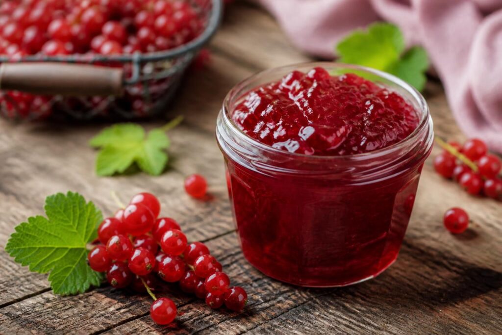 Selbst gemachtes Johannisbeergelee in einem Einmachglas, leuchtend rot und klar, auf einem Holztisch mit frischen roten Johannisbeeren daneben