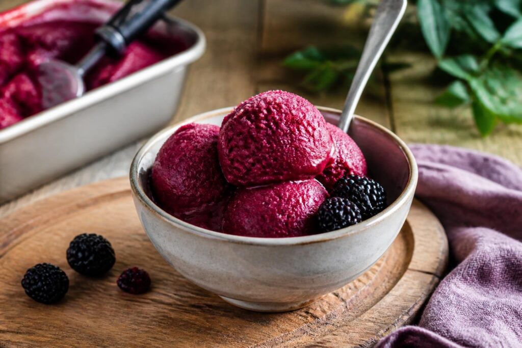Cremiges Brombeer-Sorbet in einer weißen Schale, garniert mit frischen Brombeeren und Minzblättern, auf einem hellen Holztisch