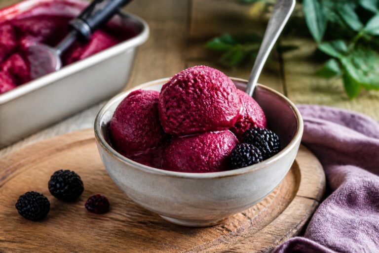 Cremiges Brombeer-Sorbet in einer weißen Schale, garniert mit frischen Brombeeren und Minzblättern, auf einem hellen Holztisch