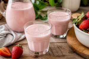 Ein hohes Glas cremige Erdbeermilch nach Omas Art, garniert mit einer frischen Erdbeere am Glasrand, auf einem Holztisch mit sommerlichem Hintergrund