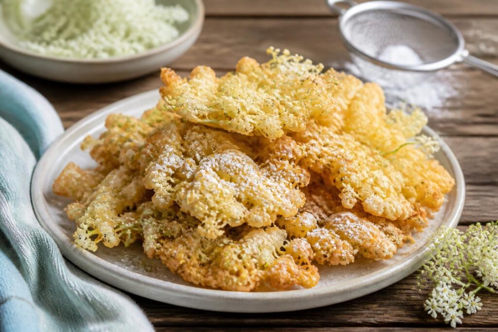 Frittierte Holunderblüten mit Puderzucker bestäubt auf einem Holzbrett, goldbraun und knusprig