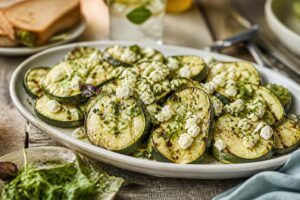 Gegrillte Zucchinistreifen mit zerbröckeltem Feta, frischen Kräutern und Zitronenabrieb auf einem Holzbrett