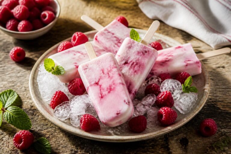 Selbst gemachte Himbeer-Joghurt-Eis am Stiel mit zweischichtigem Farbverlauf auf einem Holzbrett, frische Himbeeren daneben