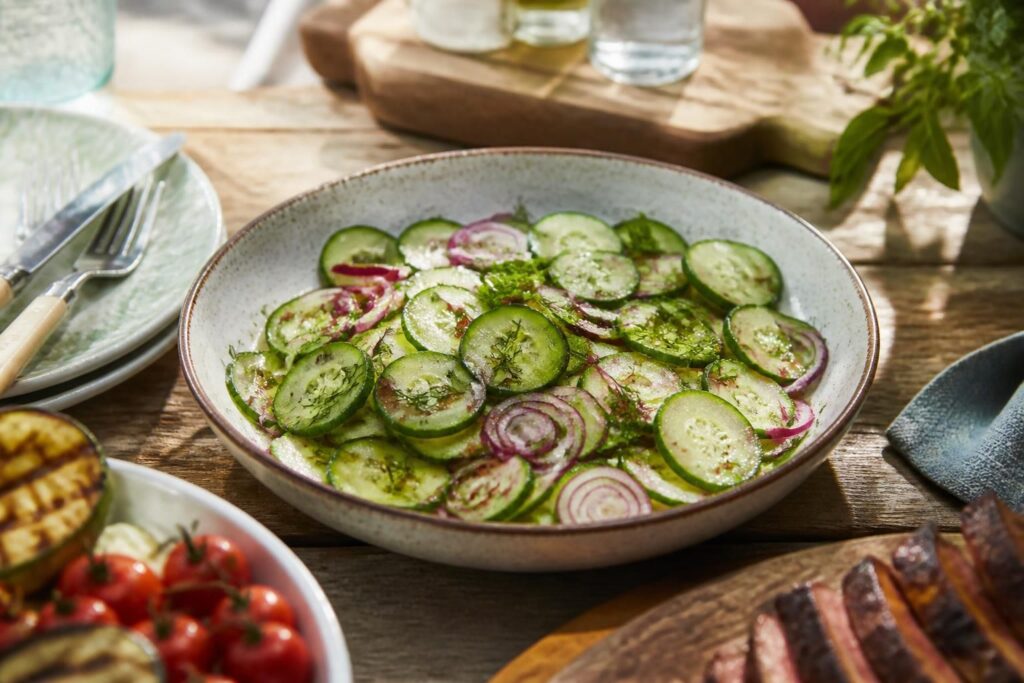 Jamie Olivers Gurkensalat mit Dill, Zitrone und Olivenöl in einer weißen Schüssel auf einem Holzbrett – perfekt fürs Grillbuffet