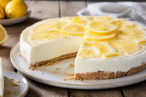 Kalte Zitronentorte ohne Backen mit Joghurtcreme ohne Gelatine, sauber angeschnitten auf einem weißen Teller, Keksboden und frische Zitronenscheiben sichtbar