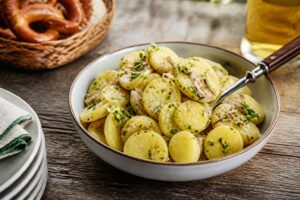 Kalter Kartoffelsalat mit Essig und Öl in einer weißen Schüssel, garniert mit Schnittlauch, serviert im Biergarten auf einem Holztisch