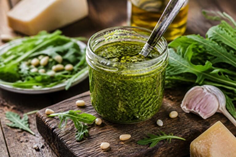 Selbstgemachtes Rucola-Pesto im Schraubglas, umgeben von frischem Rucola, Parmesan, Pinienkernen und einem Schuss Olivenöl auf einem Holzbrett