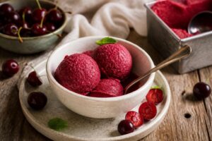Sauerkirsch-Sorbet ohne Eismaschine in einer weißen Schale, rubinrote Kugeln garniert mit frischer Minze und ganzen Sauerkirschen