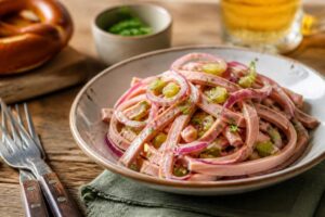 Wurstsalat mit Essig-Öl-Dressing in einer weißen Schüssel, garniert mit Schnittlauch und roten Zwiebeln, serviert mit Laugenbrezel auf einem Holzbrett
